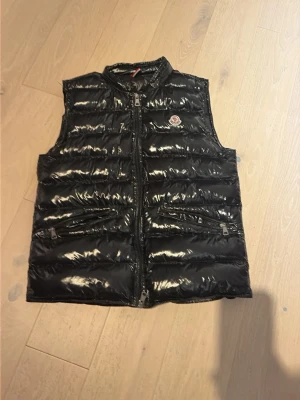 Svart glansig dunväst från Moncler - Snygg svart dunväst från Moncler Longue Saison med glansig finish och två dragkedjefickor framtill. Västen har hög krage och Moncler-logga på bröstet. Perfekt för lager-på-lager och streetstyle.