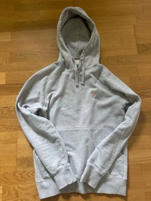 Grå hoodie från Revolution - Snygg grå hoodie från Revolution med broderad liten röd detalj på bröstet. Klassisk modell med huva och dragsko samt magficka. Tillverkad i mjukt material som känns skönt mot huden. Perfekt för en avslappnad och stilren look.