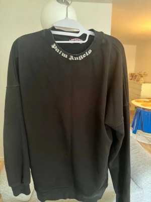 Svart sweatshirt från Palm Angels - Svart oversized sweatshirt från Palm Angels med logotyp i vitt tryckt runt halsringningen. Tröjan har långa ärmar, ribbade muddar och en avslappnad passform. Perfekt för en streetwear-look.