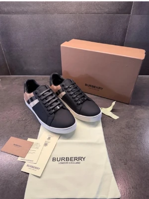 Burberry svart-beige sneakers - Snygga sneakers från Burberry i svart läder med klassiskt beige och vitt rutmönster på sidan. Skorna har vita sulor och svarta skosnören. Perfekta för dig som vill ha en stilren och lyxig look med en touch av brittisk design.