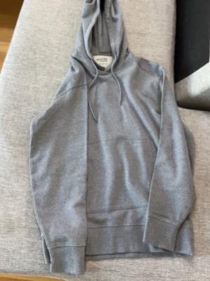 Grå hoodie från Selected Homme - Snygg grå hoodie från Selected Homme med klassisk huva och dragsnören. Tillverkad i mjukt bomullsmaterial och har ribbade muddar vid ärmslut och nederkant. Perfekt för en avslappnad och stilren look.