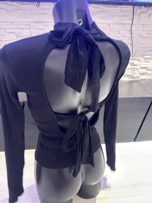 Svart ribbad topp med öppen rygg - Trendig svart ribbad topp med lång ärm och hög krage. Baksidan är helt öppen med två stora knytband i nacken och midjan för en snygg och unik look. Perfekt för dig som vill sticka ut och gillar detaljer.