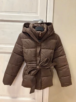 Brun pufferjacka med huva och bälte - Passar bättre på en M eller L!!!! Den var lite för trång på mig som brukar ha XL!! Snygg brun pufferjacka med huva och matchande bälte i midjan. Jackan har quiltad design, dragkedja och tryckknappar framtill. Perfekt för kalla vinterdagar och ger en trendig siluett. Tillverkad i vadderat material för extra värme.