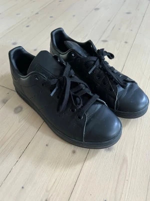 Svarta Adidas Stan Smith sneakers i storlek 38 - Säljer ett par helsvarta Adidas Stan Smith sneakers med klassisk låg profil och svarta skosnören. Skorna är tillverkade i syntetmaterial och har diskreta perforerade ränder på sidorna. Primegreen-material för en mer hållbar stil.