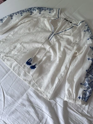 Vit blus med blå brodyr från Zara - Vit blus från Zara med blå broderade detaljer längs ärmar och halsringning. Blusen har långa ärmar och knytband med tofsar framtill. Tillverkad i ett lätt och luftigt material, perfekt för en avslappnad och trendig look.
