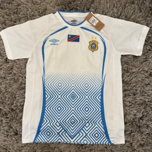 DR Kongo fotbollströja från Umbro - Säljer en vit och blå DR Kongo fotbollströja från Umbro med officiella klubbmärket och flagga på bröstet. Tröjan har korta ärmar, rund hals och ett grafiskt mönster i blått nedtill. Perfekt för match eller träning!