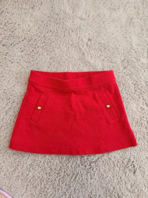 Röd kortkjol från Janie and Jack - Snygg röd kortkjol från Janie and Jack med dekorativa guldknappar framtill. Kjolen har en enkel och stilren design.  Kika in min profil för fler fynd! Öppen för prisförslag. 