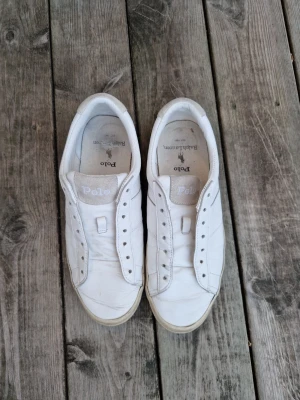 Vita sneakers från Polo Ralph Lauren - Stilrena vita sneakers från Polo Ralph Lauren i läder med klassisk låg profil och broderad logga på sidan. Skorna har vita sulor och saknar skosnören, vilket ger en clean look. Perfekta för en avslappnad och trendig stil.