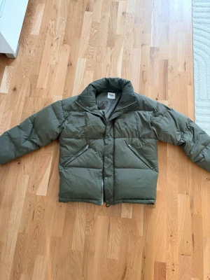 Olivgrön pufferjacka från ZARA - Snygg olivgrön pufferjacka från ZARA med hög krage och två dragkedjeförsedda fickor framtill. Jackan har en quiltad design och stängs med dragkedja. Perfekt för kalla vinterdagar och ger en trendig look.
