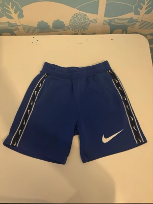 Blå Nike shorts med vita detaljer - Säljer ett par blå Nike shorts med vita loggor längs sidorna och en stor vit swoosh på ena benet. Shortsen har elastisk midja och sportig look, perfekta för träning eller chill. Materialet känns mjukt och följsamt.