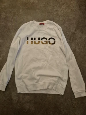 Vit sweatshirt från HUGO - Snygg vit sweatshirt från HUGO med stor logga i metallic guld och svart framtill. Tröjan har rund halsringning, långa ärmar och ribbade muddar vid ärmslut och nederkant. Perfekt för en clean och trendig look. Står storlek M men är storlek S