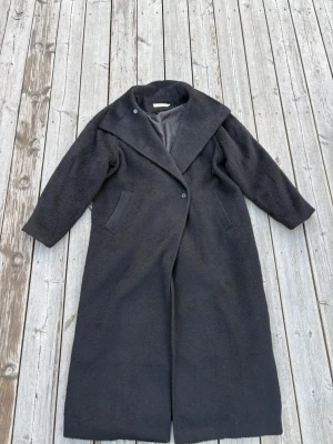 Lång svart  - Snygg lång svart kappa med knäppning framtill. Kappan har två sidofickor och en rak, avslappnad passform.  Perfekt för kyliga dagar och ger en stilren look.