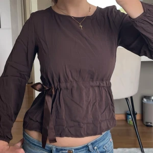 Brun blus med sidenband i midjan - Mörkbrun blus med rund halsringning och långa ärmar. Den har ett sidenband som markerar midjan och som knyts i sidan 🤎