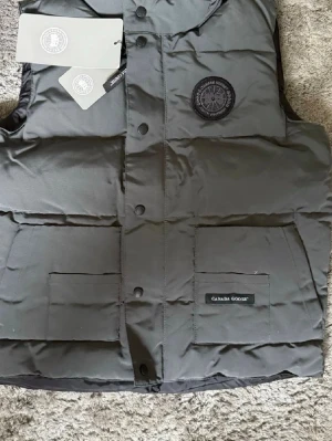 Grå dunväst från Canada Goose - Snygg grå  dunväst från Canada Goose med två stora framfickor och diskret logga på bröstet. Västen har tryckknappar framtill och är vadderad för extra värme. Perfekt för lager-på-lager och streetwear-stil.
