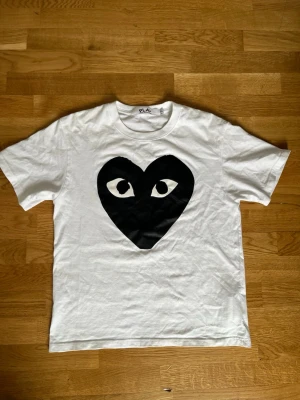 Vit Comme des Garçons Play t-shirt - Säljer en vit t-shirt från Comme des Garçons Play med det ikoniska svarta hjärtat med ögon på bröstet. Klassisk passform och rund halsringning. Perfekt för dig som gillar streetwear och vill ha något stilrent men ändå unikt.