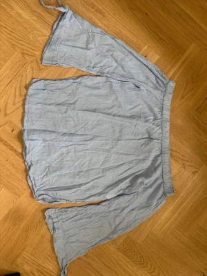 Blå offshoulder blus med ränder - Ljusblå offshoulder blus med tunna vita ränder och resår vid axlarna. Blusen har vida, långa ärmar och är gjord i ett lätt och luftigt tyg, perfekt för en avslappnad och trendig look.