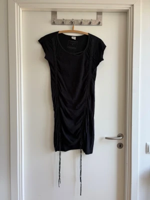 Svart draperad klänning från Object - Snygg svart klänning från Object med draperade detaljer och justerbara snören framtill. Kortärmad modell i mjukt material med rund halsringning. Perfekt för dig som gillar en avslappnad men trendig stil.