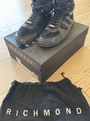 Svarta Richmond high top sneakers - Snygga svarta high top sneakers från Richmond med detaljer i mocka, lack och textil. Skorna har silverfärgade nitar på plösen och logotyp på sidan. Säljes med originalkartong och dustbag. Perfekta för dig som vill sticka ut med din stil. Nypris 3800kr köpta i Dubai