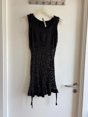 Svart ärmlös klänning från Body - Snygg svart ärmlös klänning från Body i storlek 38. Klänningen har ett unikt virkat mönster på nederdelen och knytband i sidorna. Perfekt för dig som vill ha en cool och annorlunda look.