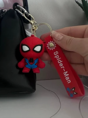 Spider-Man nyckelring med figur - Nyckelring med en röd Spider-Man figur i mjukt material och en matchande röd rem med texten 'Spider-Man' och spindelmotiv. Perfekt att hänga på väskan eller använda till nycklar. Färgerna är röd, blå, svart och vit.