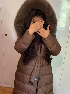 Lång brun dunjacka med pälshuva - Snygg lång brun dunjacka med stor huva kantad av fluffig fuskpäls. Jackan har bälte i midjan för en schysst siluett och quiltad design som håller värmen. Perfekt för kalla vinterdagar och riktigt mysig att bära.