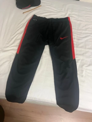 Svarta Nike mjukisbyxor med röda detaljer - Säljer ett par svarta mjukisbyxor från Nike med röda detaljer längs sidorna och röd Nike-logga på ena benet. Byxorna har dragsko i midjan och muddar vid bensluten. Perfekta för chill eller träning.