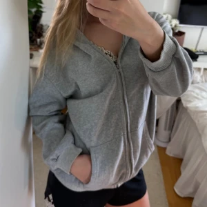 Grå zip hoodie med huva - Säljer en enkel och stilren grå hoodie med dragkedja och huva. Hoodien har två fickor framtill och ribbade muddar vid ärmsluten. Perfekt för chill dagar eller när du vill ha en avslappnad look.