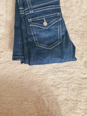 Mörkblå jeans med kontrastsömmar - Snygga mörkblå jeans med vita kontrastsömmar och klassisk femficksdesign. Jeansen har en bakficka med lock och knappdetalj samt låg midja och utsvängda ben💕 strl 152
