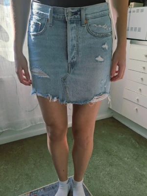 Ljusblå Levi's jeanskjol med slitningar W27 - Trendig ljusblå jeanskjol i kort modell med rå kant och slitna detaljer. Kjolen har klassiska fem fickor, bälteshällor och knappgylf. Perfekt för en avslappnad och cool stil.