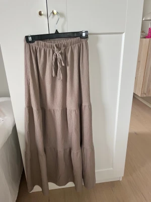 Beige långkjol med knyt i midjan - Säljer en beige långkjol med resår och knytband i midjan. Kjolen har en luftig och flowy passform med volangdetaljer och är tillverkad i ett mjukt, strukturerat tyg. Perfekt för en avslappnad och trendig look.