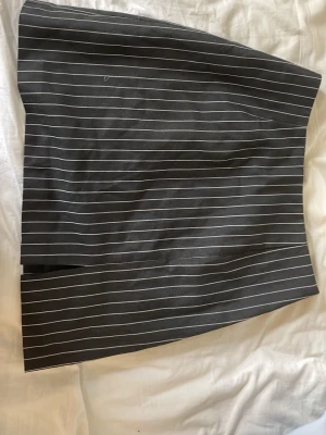 Mörkgrå kortkjol med vita ränder - Snygg mörkgrå kortkjol med tunna vita ränder och en liten slits framtill. Kjolen har en rak passform och är perfekt för en stilren och trendig look. Passar till både vardag och mer uppklädda tillfällen.