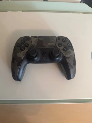 Kamouflagefärgad PS5 handkontroll - LÄS INNAN DU KÖPER: DENNA KONTROLL HAR STICKDRIFT MEN OM DU VET HUR MAN FIXAR DET SÅ ÄR DEN PERFEKT FÖR JUST DIG. Snygg handkontroll till PlayStation 5 med grått och svart kamouflagemönster. Ergonomisk design med två analoga spakar, styrkors och klassiska PlayStation-knappar. Tillverkad i plast och passar perfekt för gaming.