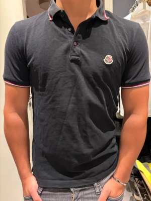 Svart pikétröja från Moncler - Klassisk svart pikétröja från Moncler med korta ärmar och knappslå framtil. Jag är 177 cm lång och väger 76 kg. Tveka inte på höra av dig vid dm. 💪💪