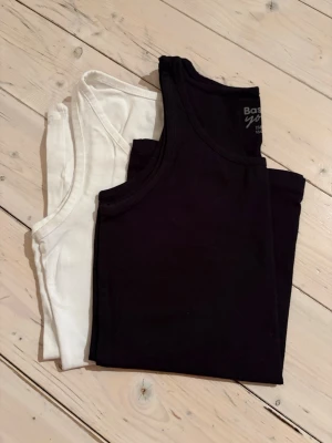 Svart och vit linnetopp i bomull - Två klassiska linnetoppar i svart och vitt. Båda är ärmlösa och har rund halsringning. Perfekta att styla lager på lager eller bära som de är. Tillverkade i mjuk bomull för maximal komfort.
