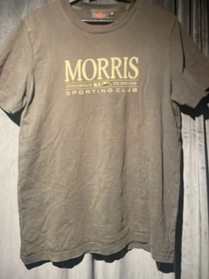 Grå t-shirt från Morris - Säljer en grå t-shirt från Morris med tryck 'Morris Sporting Club' i gult på bröstet. Klassisk rund hals och korta ärmar. Perfekt till jeans eller shorts för en avslappnad stil.