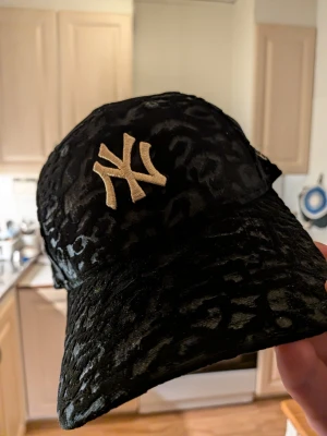 Svart New Era NY keps i sammet - Säljer en svart keps från New Era med leopardmönster i sammet och guldfärgad NY-logga framtill. Kepsen har böjd skärm och broderad logga på sidan. Perfekt för dig som vill sticka ut med en unik accessoar. Helt ny. 