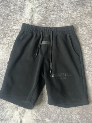 Svarta shorts från Fear of God Essentials - Svarta shorts från Fear of God Essentials med snörning i midjan och diskret logga på vänster ben. Tillverkade i mjuk bomull med normal passform och två sidofickor. Perfekta för en avslappnad och stilren look.
