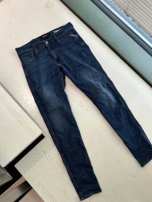 Mörkblå Replay Bronny jeans - Snygga mörkblå jeans från Replay, modell Bronny Super Slim Fit. Jeansen har klassisk femficksdesign, dragkedja och knapp framtill samt Replay-detaljer på fickan. Perfekta för dig som gillar en smal passform och stilren look.