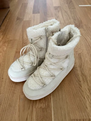 Vita fluffiga moon boots med snörning oanvända - Säljer ett par vita moon boots med fluffigt foder och grov snörning framtill. Bootsens ovandel är i fårskinnsimitation och de har dragkedja på sidan för enkel påtagning. Perfekta för kalla dagar när du vill hålla värmen och ändå se stylish ut. Något mindre i storlek.
