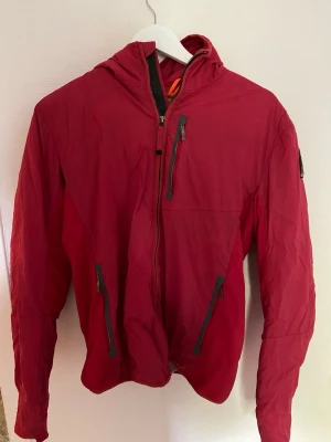 Röd vindjacka från Parajumpers - Snygg röd vindjacka från Parajumpers med huva och dragkedja framtill. Jackan har tre fickor med dragkedja och ett tydligt PJS-märke på ärmen. Lätt material som passar perfekt till blåsiga dagar. Sportig och modern design.