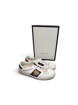 Gucci vita sneakers med bi-detalj - Snygga vita sneakers från Gucci med klassiska gröna och röda sidoband samt guldbroderad bi. Skorna har vita snören, låg profil och detaljer i grönt och rött på hälen. Tillverkade i läder för en lyxig känsla och stilren look. Pris kan diskuteras. Snabb frakt📦