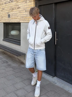 Ljusgrå zip hoodie från parajumpers - Snygg ljusgrå hoodie med dragkedja och huva. Har två stora fickor framtill och en patch på vänster ärm. Justerbara snören i huvan och ribbade muddar vid ärmslut och nederkant. Perfekt till jeans eller shorts för en avslappnad look.