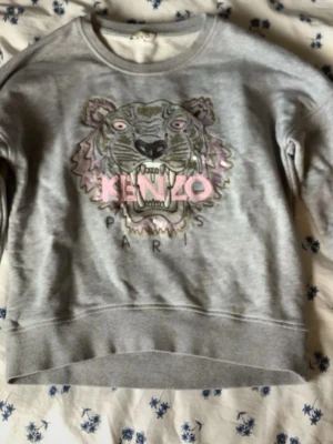 Grå sweatshirt med tigertryck från Kenzo - Säljer en grå sweatshirt från Kenzo med det ikoniska tigerhuvudet broderat framtill i rosa, lila och svart. Tröjan har rund halsringning och ribbade muddar vid ärmslut och nederkant. Perfekt för dig som gillar streetwear och vill sticka ut.