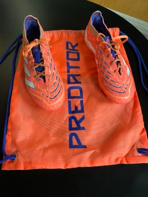 Fotbollsskor - Säljer ett par grymma Adidas Predator fotbollsskor i orange med blå och vita detaljer Kommer med matchande orange påse. Storlek 38.2/3 