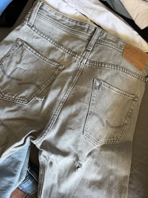 Grå jeans från Jack & Jones - Snygga grå jeans från Jack & Jones i modellen Relaxed Chris. Jeansen har klassisk femficksdesign, raka ben och knäppning med knappar. Tillverkade i bomull med en cool tvättad look och detaljerade bakfickor. 2 st hål där bak på dom därför billigt. 