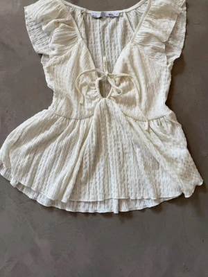 Vit volangblus med knytning - Söt vit blus i lätt, krinklat tyg med volangärmar och v-ringning med knytning framtill. Blusen har en luftig passform och markerad midja som ger en feminin siluett. Perfekt till jeans eller kjol för en trendig look.