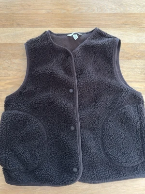 Mörkbrun pileväst med stora fickor - Mysig mörkbrun pileväst från H&M i fluffigt material med rund halsringning och tryckknappar framtill. Västen har två stora runda fickor och kantband i matchande färg. Perfekt för lager på lager och en chill look.