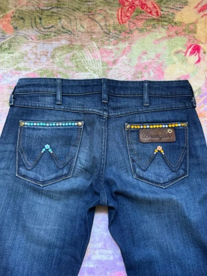 Snygga blåa lågmidjade jeans med pärldetaljer - Unika Mörkblå bootcut jeans med låg midja från vintage Wrangler😍De har dekorativa pärlor, blommor och stenar i blått och orange på bakfickorna. Så snyggt som verkligen gör din outfit så coolare🩷Klassisk femficksmodell med bälteshällor och snygga kontrastsömmar. Framsidan har färgade snäckor, pärlor och samma stenar som en blomma🩵Materialet är denim i bomull med en mjuk tvättad finish! Storlek XS, W26/L32 i modellen Megan. Mått cm: midja 36, gren 19, innerben 80. Använda men inga defekter❤️