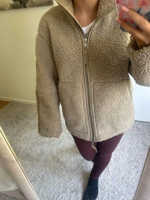 teddyjacka  - Mysig beige brun teddyjacka med hög krage och grov dragkedja framtill. Jackan har två stora fickor på sidorna och är tillverkad i fluffigt, mjukt teddy-material. Perfekt för kalla dagar när du vill vara både varm och stilren.