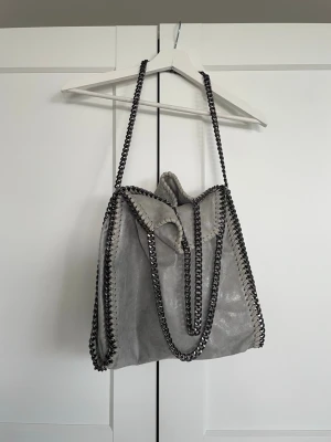 Grå tiamo axelremsväska med kedjedetaljer - Snygg grå handväska i mjukt material med silverfärgade kedjor längs kanterna och som axelremmar. Väskan är rymlig och liknar Stella McCartney, den har även flera innerfack och med en djurmönstrad insida. I mycket fint skick och knappt använd. Säljer då den inte kommer till användning. 